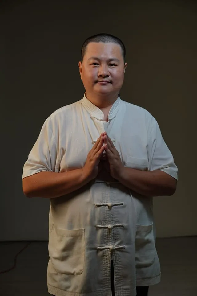 緣起順周 2 這是一張專業整復師的形象照,穿著中式唐裝上衣、雙手合十,背景簡潔、光影聚焦臉部與上半身,整體呈現出穩重、尊敬的風格