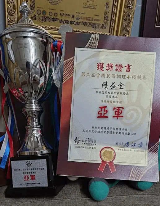 陳益全師傅得過第二屆全國民俗調理手技競賽的獎狀與獎盃，由社團法人民俗調理業從業技能發展協會的唐江雲理事長所頒發